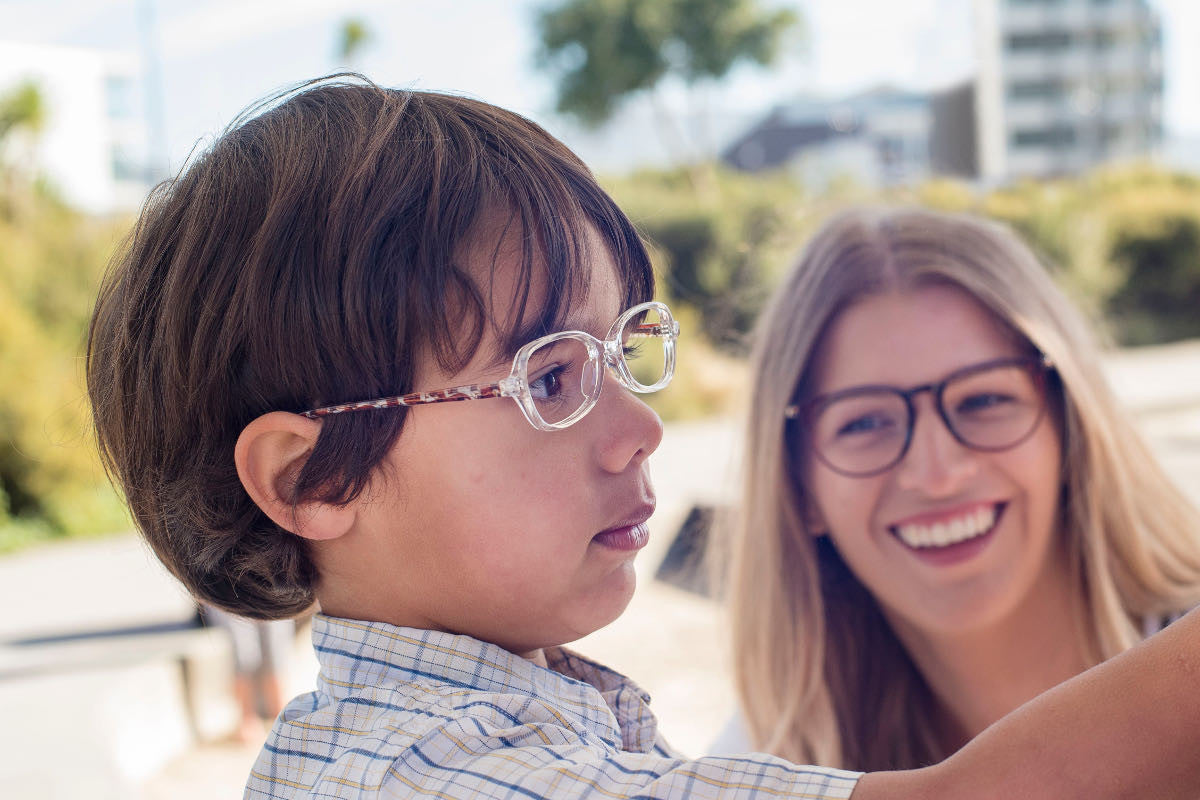 Child wearing clear acetate children's glasses frames by Mr Foureyes, standing at a blackboard, with a woman wearing glasses smiling at him